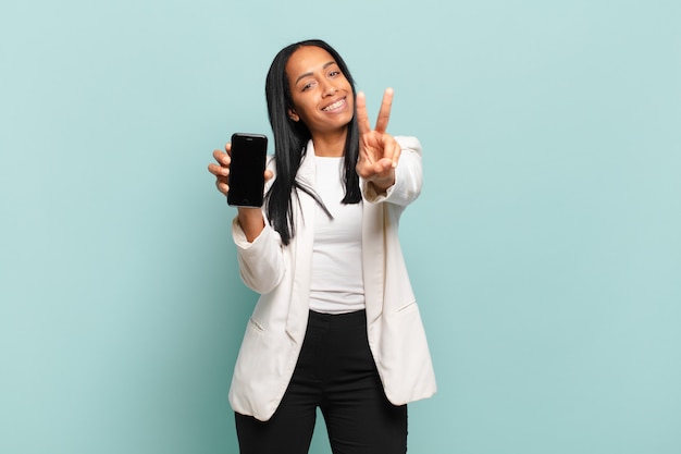 Junge schwarze Frau, die lächelt und glücklich, sorglos und positiv schaut, Sieg oder Frieden mit einer Hand gestikulierend. Smartphone-Konzept