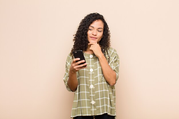 Junge schwarze Frau, die mit einem glücklichen, selbstbewussten Ausdruck mit der Hand am Kinn lächelt, sich wundert und mit einem Smartphone zur Seite schaut