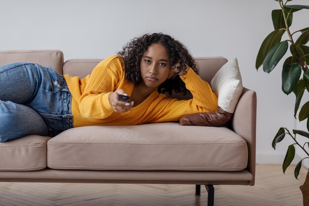 Junge schwarze Frau mit Fernbedienung, die auf dem Sofa liegt und sich gelangweilt beim Fernsehen umschaltet