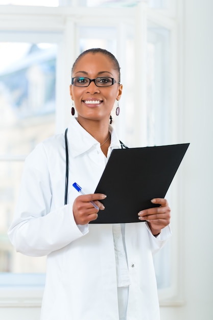 Junge schwarze Ärztin, die an einem Fenster in der Klinik mit einer Akte oder einem Dossier steht