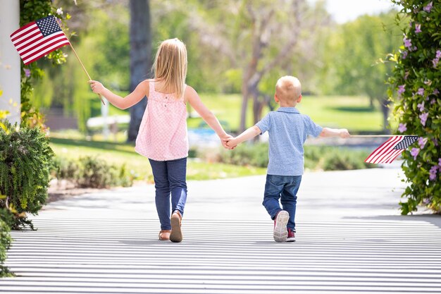 Junge Schwester und Bruder halten sich an den Händen und schwenken amerikanische Fahnen im Park