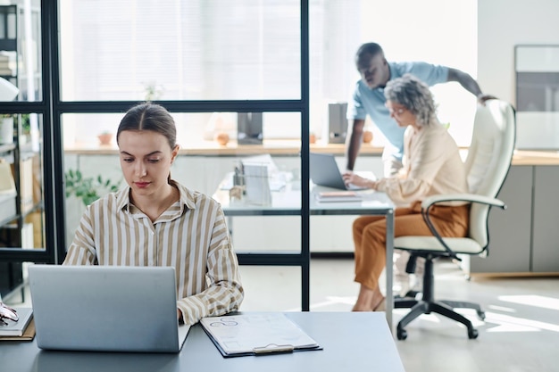 Junge Sekretärin oder Analytikerin benutzt Laptop, während sie mit Dokumenten am Schreibtisch sitzt