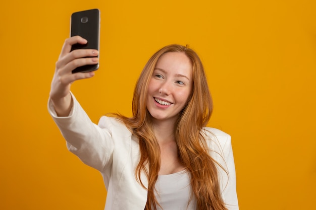 Junge, selbstbewusste, erfolgreiche und schöne geschäftliche rothaarige Frau, die selfie mit dem Smartphone auf gelb nimmt. Beruf, Karriere, Berufsbild.