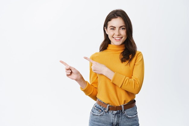 Junge selbstbewusste Frau, die Logo auf leerem Raum zeigt Lächelndes Mädchen, das einlädt oder Werbeaktionen anbietet, die mit dem Finger nach links zeigen und auf den weißen Hintergrund der Kamera schauen