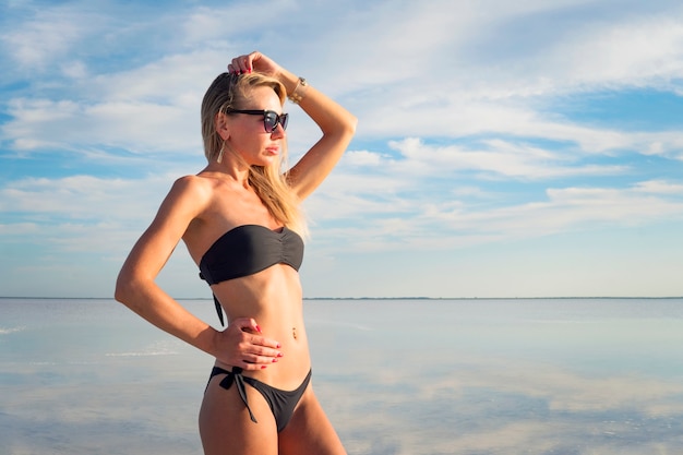 Junge sexy Dame mit dem schwarzen Badeanzug im Urlaub. Model posiert in der Natur. Mädchen, das in die Ferne im Hintergrundwasser mit Reflexionen des bewölkten Himmels schaut