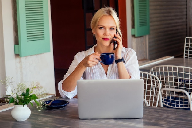 Foto junge sexy frau in einem stylischen hemd mit laptop und telefon freiberuflich am tropischen paradiesstrand mädchen tasse tee kaffee freiberuflerin arbeit sitzend in einem sommercafé fernarbeit indischer ozean