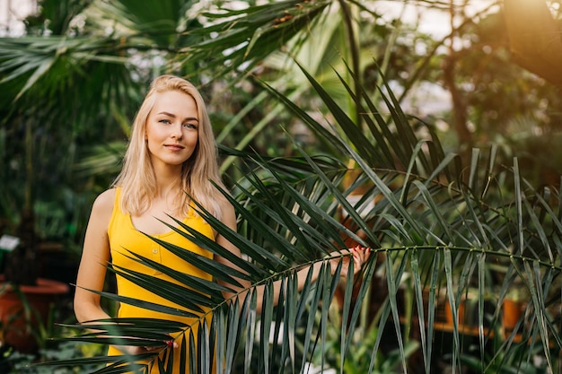 Junge sexy Frau mit schlankem Körper, tragenden gelben Badeanzug, der unter tropischen Bäumen steht. Horizontale Aufnahme eines charmanten Mädchens, das während des Urlaubs im Resort in grünen Blättern posiert. Sommerurlaub.