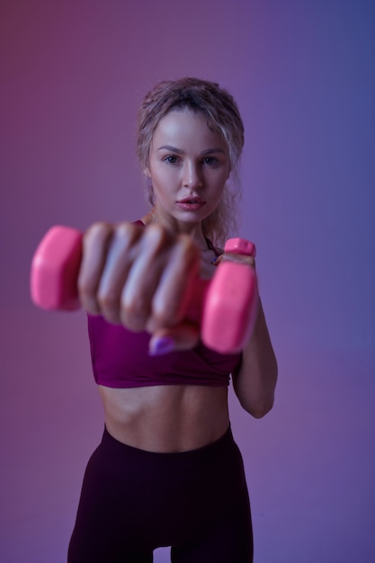 Junge sexy Sportlerin mit Hanteln posiert im Studio, Neonhintergrund