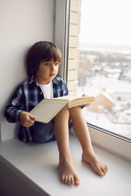 Junge sitzt auf dem Fenster eines Hauses mit einem Buch im Winter auf einer hohen Etage