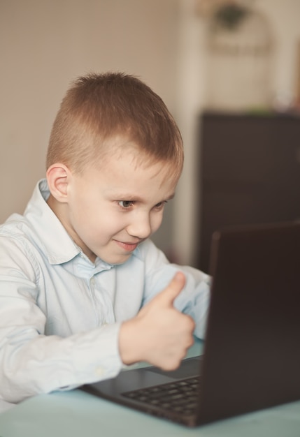 Junge sitzt auf dem Tisch und spricht online. Arbeitsplatz Schreibtisch mit Laptop. Kind lernt online. Online-Fernunterricht.