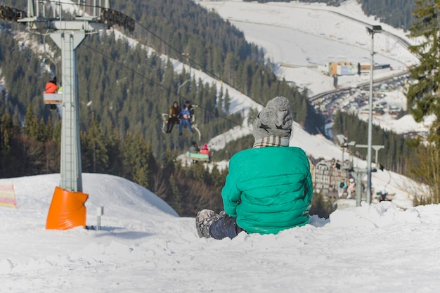 Foto junge sitzt auf einem schneebedeckten hügel auf dem hintergrund des pinienwaldes der seilbahn und der berge skigebiet wintertag