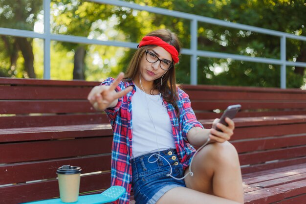 Junge Skaterin in stilvollen Kleidern sitzt auf der Tribüne in einem Skatepark, hält ein Smartphone in den Händen und hört an einem strahlend sonnigen Tag Musik in Kopfhörern