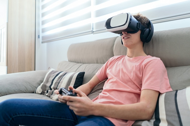 Junge spielt Konsole mit Virtual-Reality-Brille-Kopfhörern und Joystickin im Wohnzimmer