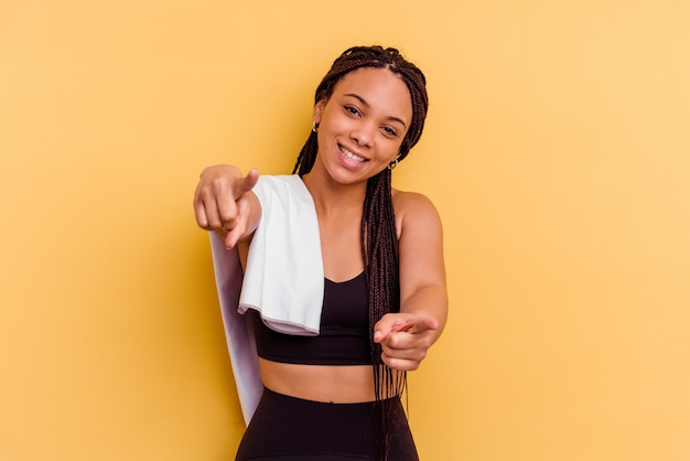 Junge Sport Afroamerikanerfrau, die ein Handtuch lokalisiert auf gelbe Wand fröhliches Lächeln hält, das nach vorne zeigt.