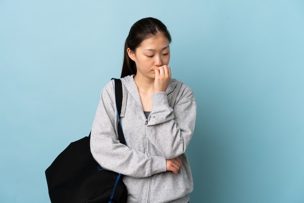 Junge Sport-Chinesin mit Sporttasche über blauer Wand, die Zweifel hat