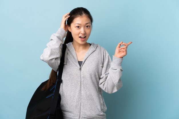 Junge Sport-Chinesin mit Sporttasche über isolierter blauer Wand überrascht und Finger zur Seite zeigend