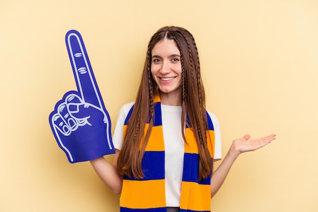 Junge Sportfanfrau isoliert auf gelbem Hintergrund, die einen Kopierraum auf einer Handfläche zeigt und eine andere Hand an der Taille hält.