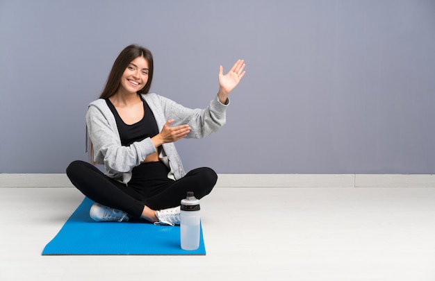 Junge Sportfrau, die auf dem Boden mit den ausdehnenden Händen der Matte zur Seite sitzt, damit die Einladung kommt