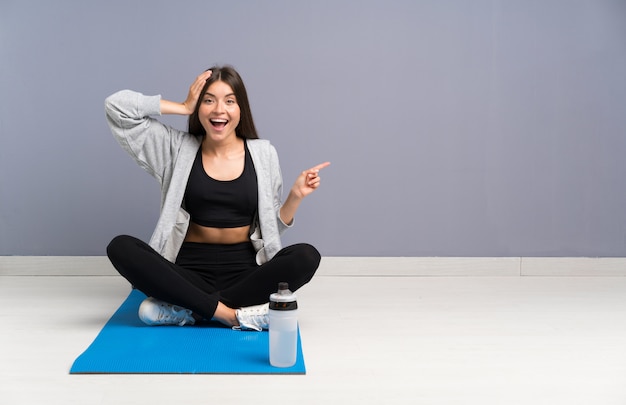 Junge Sportfrau, die auf dem Boden mit der Matte überrascht sitzt und Finger auf die Seite zeigt