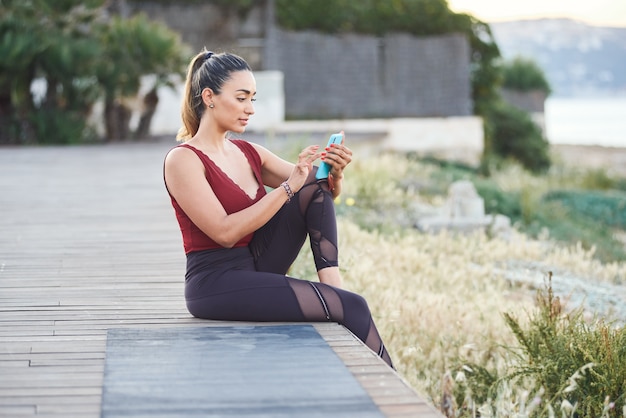 Junge Sportfrau, die mit Handy am felsigen Strand ruht.