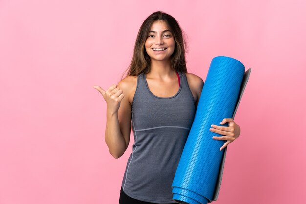 Junge Sportfrau, die zu Yoga-Klassen geht, während eine Matte lokalisiert auf rosa Wand hält, die zur Seite zeigt, um ein Produkt zu präsentieren