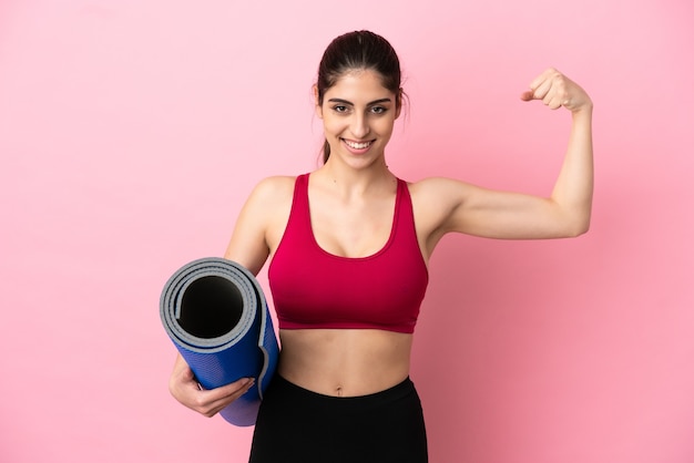 Junge sportkaukasische Frau, die zu Yoga-Kursen geht, während sie eine Matte hält und eine starke Geste macht