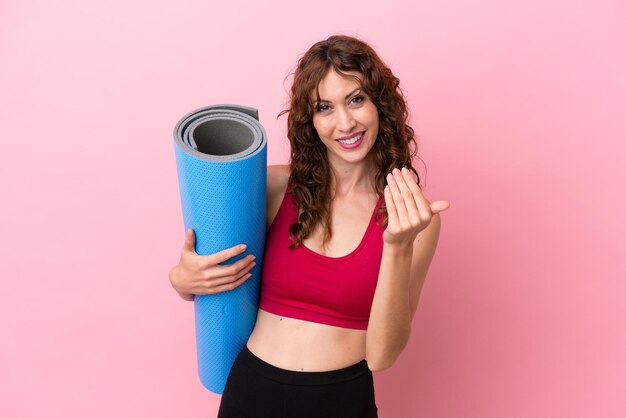 Junge Sportlerin, die zu Yoga-Kursen geht, während sie eine Matte isoliert auf rosafarbenem Hintergrund hält und einlädt, mit der Hand zu kommen. Schön, dass du gekommen bist