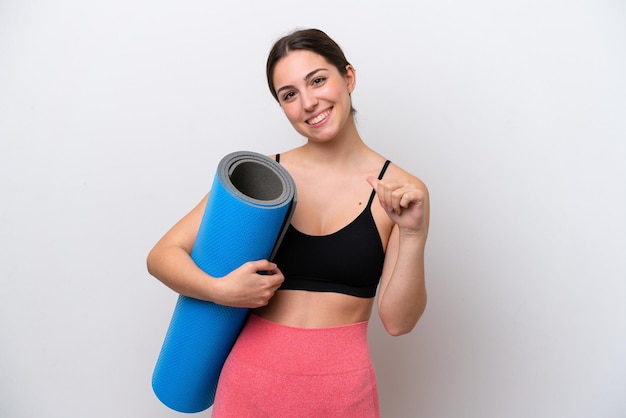 Junge Sportlerin, die zu Yoga-Kursen geht, während sie eine Matte isoliert auf weißem Hintergrund hält, stolz und selbstzufrieden