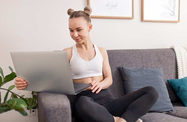 Junge Sportlerin in Sportbekleidung, die auf der Couch sitzt und Laptop durchsucht, während sie nach dem Fitnesstraining zu Hause während der Selbstisolation ruht