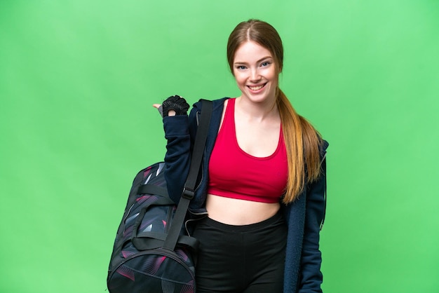 Junge Sportlerin mit Sporttasche über isoliertem Chroma-Key-Hintergrund, die zur Seite zeigt, um ein Produkt zu präsentieren