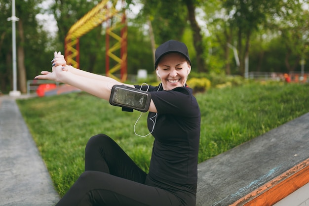 Junge sportlich lächelnde Frau in schwarzer Uniform mit Kopfhörern, die Musik hören, die Hände strecken, sich vor oder nach dem Laufen ausruhen und sitzen, im Stadtpark im Freien trainieren