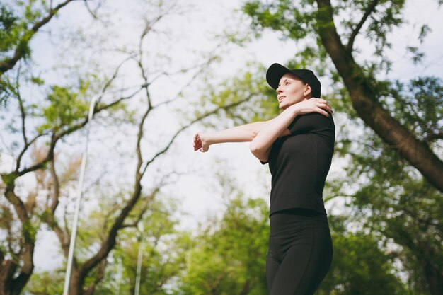 Junge sportlich schöne brünette Frau in schwarzer Uniform, Mütze, die Sportdehnungsübungen macht, Aufwärmen vor dem Laufen oder Training, im Stadtpark im Freien stehend