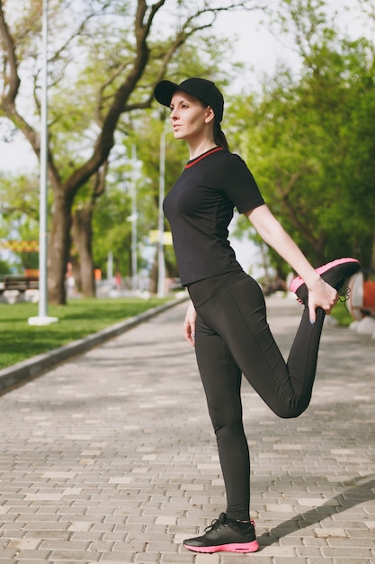 Junge sportlich schöne brünette Frau in schwarzer Uniform, Mütze, die Sportdehnungsübungen macht, Aufwärmen vor dem Laufen oder Training, im Stadtpark im Freien stehend