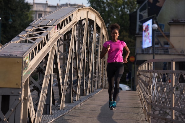 Junge sportliche afrikanisch-amerikanische Frau, die am frühen Morgen auf dem Bürgersteig über die Brücke läuft und mit der Sonnenaufgangsszene der Stadt im Hintergrund joggt