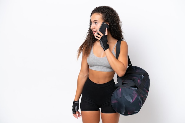 Junge sportliche arabische Frau mit Sporttasche isoliert auf weißem Hintergrund, die mit jemandem ein Gespräch mit dem Mobiltelefon führt