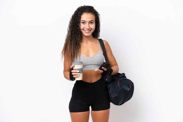 Junge sportliche arabische Frau mit Sporttasche isoliert auf weißem Hintergrund mit Kaffee zum Mitnehmen und einem Handy