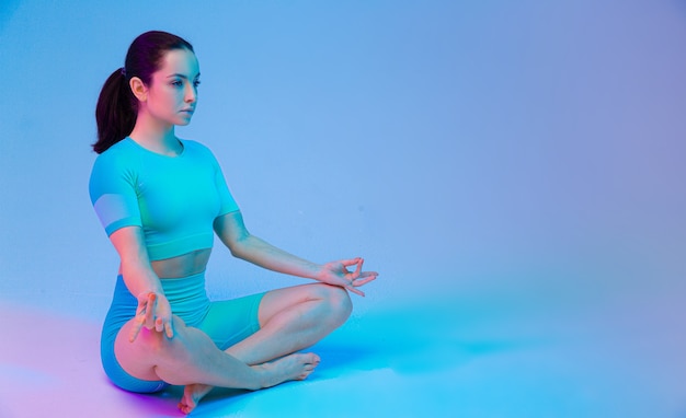 Junge sportliche attraktive Frau, die Yoga praktiziert, das in Lotus Asana sitzt, Sportkleidung, Hosen und Top trägt, weißes Yoga einzeln auf Gradientenstudiohintergrund im Neonlicht.