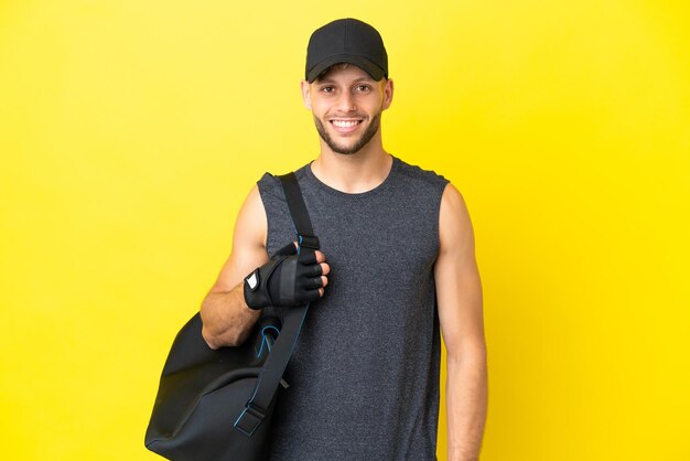 Junge sportliche blonde Mann mit Sporttasche isoliert auf gelbem Hintergrund lachen