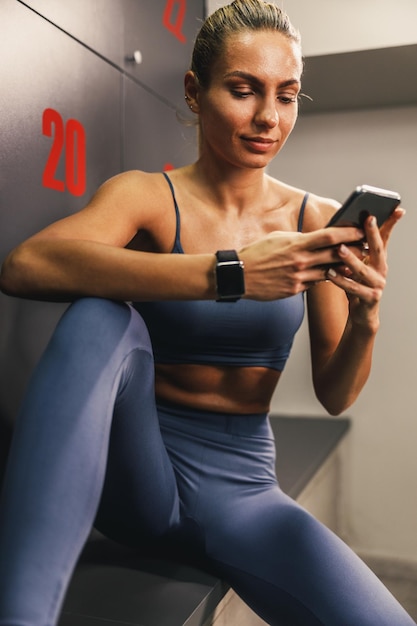 Junge sportliche Frau, die auf einer Bank in der Umkleidekabine sitzt und ein Smartphone benutzt.