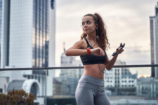 Foto junge sportliche frau, die ihre trainingsergebnisse auf der sportuhr überprüft, die mit telefon steht und musik hört