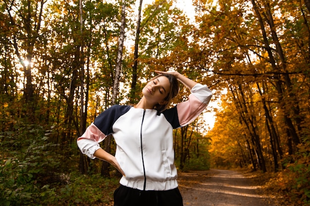 Junge sportliche Frau, die im Wald ausdehnt. Gesundes Lebensstilkonzept. Attraktive Frau mit Pferdeschwanz im Trainingsanzug, die sich vor dem Joggen aufwärmt. Fitnesskonzept im Freien. Herbstpark im Hintergrund.