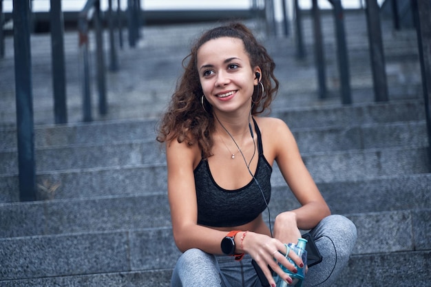 Junge sportliche Frau in Sportkleidung, die lächelt und Musik hört, während sie auf der Treppe des Stadtstadions sitzt?