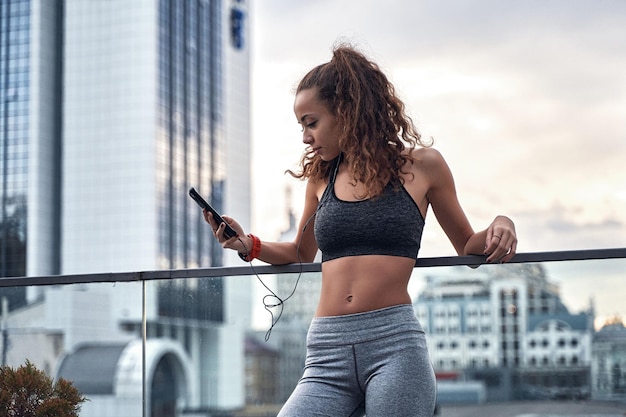 Junge sportliche Frau in Sportkleidung, die mit dem Telefon in den Händen steht, ihre Trainingsergebnisse veröffentlicht und Musik hört