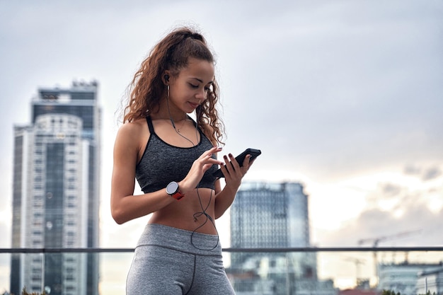 Junge sportliche Frau in Sportkleidung, die mit dem Telefon in den Händen steht, ihre Trainingsergebnisse veröffentlicht und Musik hört