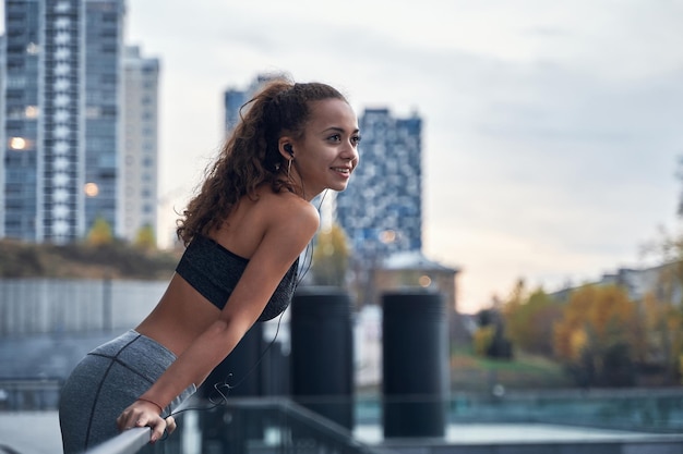 Junge sportliche Frau lehnte sich an das Geländer und hörte Musik, während sie sich ausruhte, nachdem sie auf dem Hintergrund der Stadt trainiert hatte