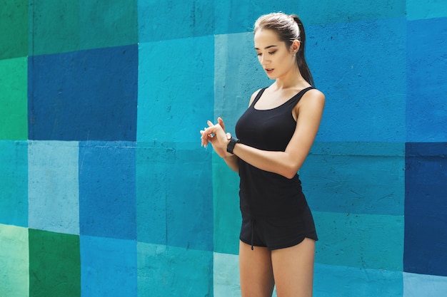 Junge sportliche Frau mit intelligenter Uhr, stehend auf hellblauem Graffiti-Wandhintergrund, Kopierraum. Modernes Technologie- und Fitnesskonzept