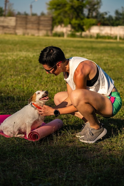 Junge sportliche Frau mit kurzen schwarzen Haaren trainiert im Freien auf Yogamatte auf grünem Gras in einem Park, Aktivität mit Hund, Jack Russell Terrier Walk