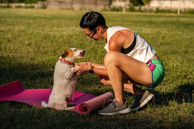 Junge sportliche Frau mit kurzen schwarzen Haaren trainiert im Freien auf Yogamatte auf grünem Gras in einem Park, Aktivität mit Hund, Jack Russell Terrier Walk