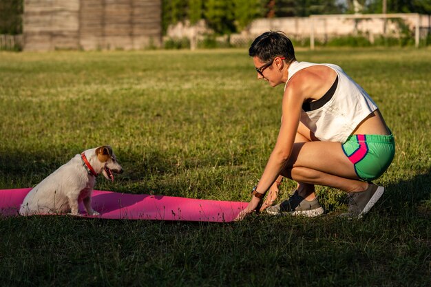 Junge sportliche Frau mit kurzen schwarzen Haaren trainiert im Freien auf Yogamatte auf grünem Gras in einem Park, Aktivität mit Hund, Jack Russell Terrier Walk