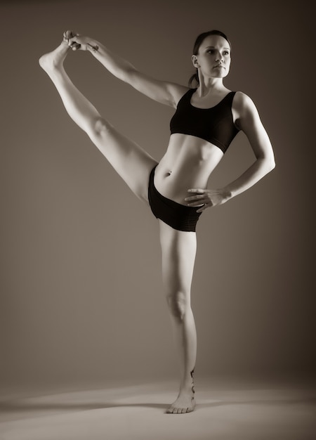 Junge sportliche Frau mit schöner Figur in schwarzer Unterwäsche macht eine Dehnung, während sie auf einem Bein auf dunklem Hintergrund steht. Konzept eines flexiblen Körpers und gesunder Gelenke. Platz für Werbung